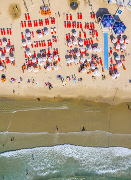 Aerial view of Tel Aviv Beach with rows of red sun loungers and white umbrellas on the sand, people relaxing near the shoreline, and gentle waves in Tel Aviv-Yafo, Tel Aviv District, Israel.