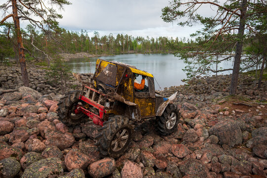 View of a muddy yellow off-road vehicle navigating over large boulders near the shore of Lake Ladoga in a forest setting in Sortavala, Respublika Kareliya, Russia.