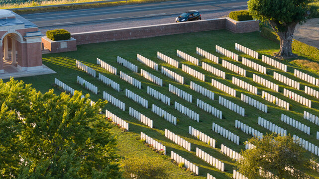 Aerial view of Warlencourt British Cemetery with rows of white headstones in green lawns, a brick entrance structure, and trees in Warlencourt-Eaucourt, Hauts-de-France, France.