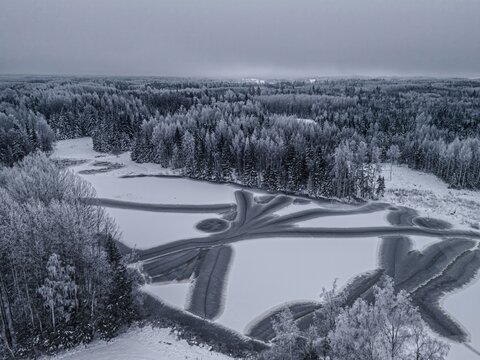 Aerial view of a frozen lake with intricate dark patterns on the ice surface surrounded by a dense snow-covered forest in Riga, Latvia.