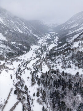 Aerial view of the snow-covered valley featuring a winding river, dense pine forests, and rugged mountain slopes under a cloudy winter sky in Val d'Aran, Catalonia, Spain.