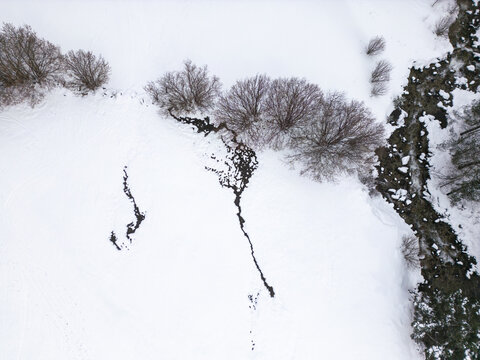 Aerial view of a snow-covered landscape featuring a winding stream and a row of bare trees in Val d'Aran, Catalonia, Spain.