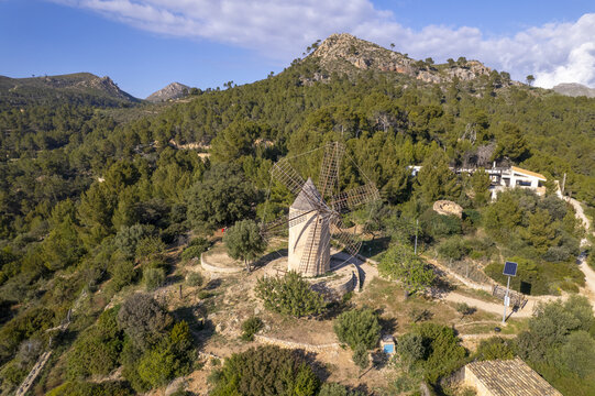 Aerial view of the historic Molino de Sa Planeta windmill situated on a pine-covered hillside with rugged mountains in the background in Andratx, Balearic Islands, Spain.