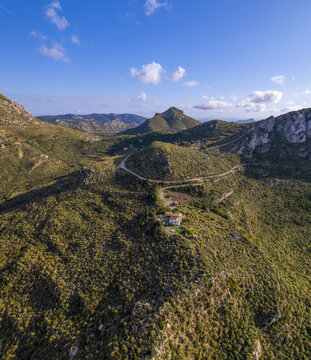 Aerial view of Coll de sa Gran Mola featuring a winding mountain road and a small stone building nestled in the rugged, green landscape in Illes Balears, Spain.