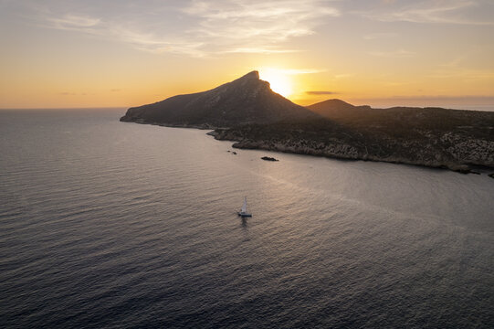 Aerial view of the rocky coastline of Sa Dragonera island at sunset with a small sailboat navigating the calm Mediterranean sea off the coast of Islas Baleares, Spain.