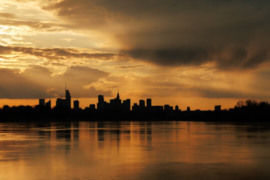 View of the Warsaw city skyline silhouetted against a dramatic golden sunset with reflections on the Vistula River, Warsaw, Masovian Voivodeship, Poland.