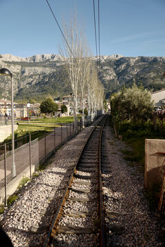 Port de Soller, Illes Balears, Spain - 01 April 2018: View of the tram tracks leading towards the Estacio de Tramvia, framed by trees with mountains in the background under a clear sky.