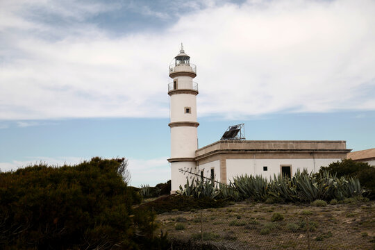 View of the historic lighthouse surrounded by agave plants and Mediterranean shrubs under a cloudy blue sky in Mallorca, Balearic Islands, Spain.