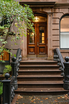 A detailed view of a classic New York brownstone entrance with carved wooden double doors and black iron railings along the steps. Green tree branches frame the historic residential facade in an urban