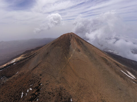 Aerial view of the volcanic peak of Mount Teide in Teide National Park with rocky slopes and clouds in the sky in La Orotava, Canary Islands, Spain.