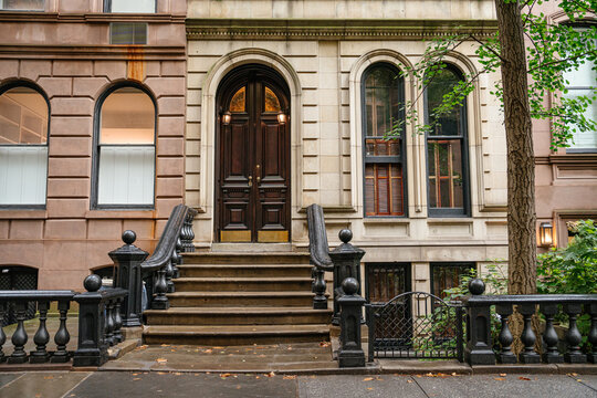 A historic New York townhouse facade with an arched wooden double door, stone exterior, and black iron staircase railings. The residential building is framed by trees on a quiet urban street.