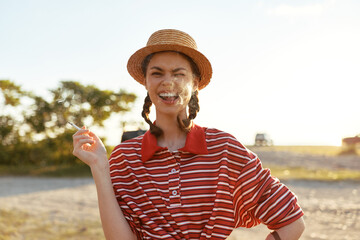 Obraz premium A joyful young woman with braided hair and a straw hat smokes a cigarette outdoors. The authentic lifestyle scene captures a relaxed moment of freedom in a sunny summer, creating a vibrant mood.