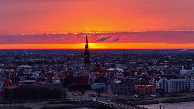 Aerial view of the historic St. Peter's Church spire rising above the rooftops of Riga at sunset, with a vibrant orange sky over the city of Riga, Latvia.