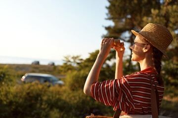 Obraz premium A woman in a striped shirt and straw hat stands outdoors during golden hour, using binoculars to survey the countryside, a moment of travel, curiosity, and lifestyle exploration.