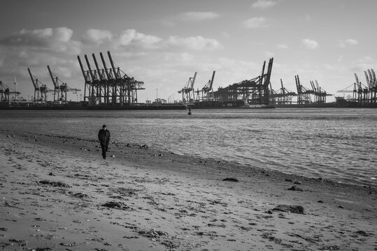 Hamburg, Germany - 14 July 2026: View of a person walking along the sandy riverbank with the industrial container cranes of the Port of Hamburg in the background.
