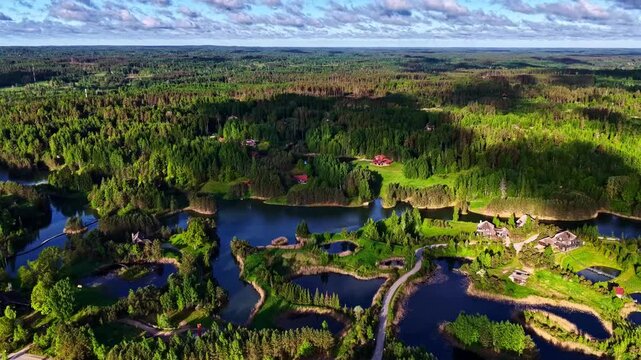 Aerial view of Amatciems village with wooden houses nestled in a lush green forest landscape surrounded by interconnected ponds and winding paths in Amatciems, Cesu novads, Latvia.