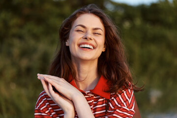Obraz premium A cheerful woman outdoors in a red striped shirt smiles with eyes closed, enjoying a sunny moment. A candid lifestyle scene highlighting happiness, relaxation, and natural joy.
