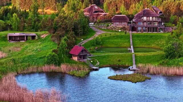 Aerial view of luxury wooden houses with thatched roofs nestled in the lush green landscape of the Amatciems eco-village with calm ponds and reeds in Amatciems, Cesu novads, Latvia.