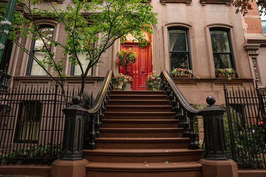 Wide view of a historic New York brownstone townhouse featuring stoop stairs, red door, black iron fence, and stone facade. A leafy tree and flower boxes enhance the urban residential streetscape.