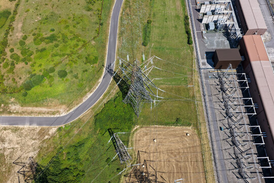 Aerial view of an electrical substation with high voltage power lines and metal pylons situated in a rural landscape with a winding road and green fields in the United Kingdom.