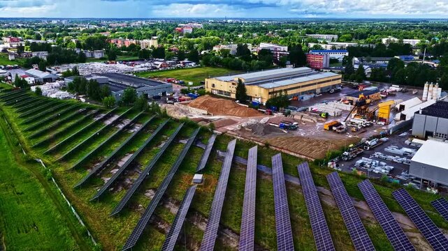 Aerial view of a solar farm with rows of photovoltaic panels adjacent to an industrial facility with warehouses and construction materials in Jelgava, Jelgava City, Latvia.