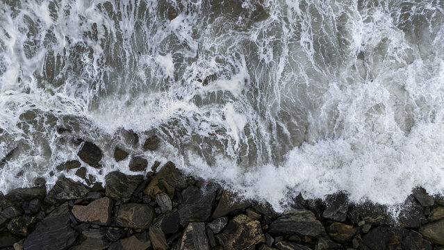 Aerial view of ocean waves crashing against dark rocky shoreline in Cape Coast, Central Region, Ghana.