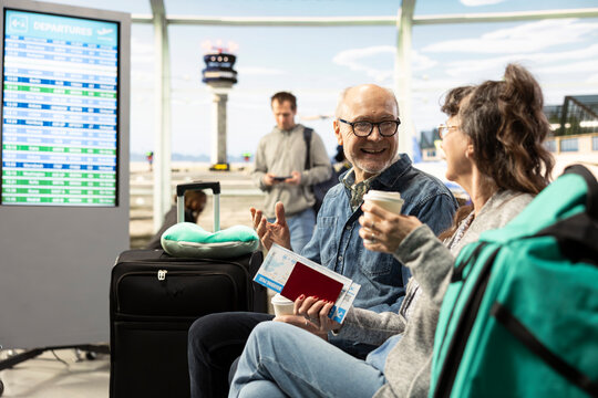 Joyful retired couple drinks coffee in an airport waiting area, discussing travel plans before departure and enjoying freedom in later life.