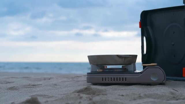 Portable camping stove and pan on sandy beach with ocean view. Portable camping stove sitting on sandy beach with pan, resting near small table and overlooking serene ocean horizon under cloudy sky
