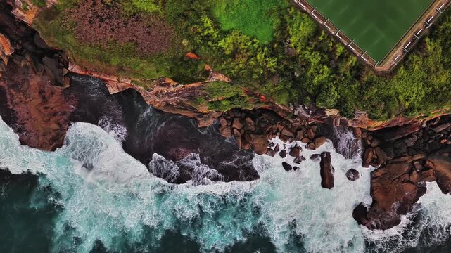 Aerial view of Bronte Baths and rocky coastline with crashing waves and green grass in Waverley, New South Wales, Australia.