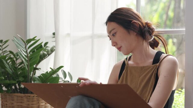 Creative young Asian woman artist painting on canvas in bright home studio. Relaxed female hobbyist sitting on floor, enjoying peaceful indoor leisure time and expressing imagination.