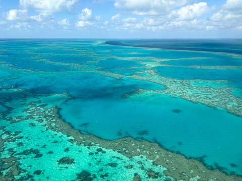 Aerial view of the Great Barrier Reef featuring vibrant coral formations and turquoise waters under a scattered cloud sky in Airlie Beach, Queensland, Australia.