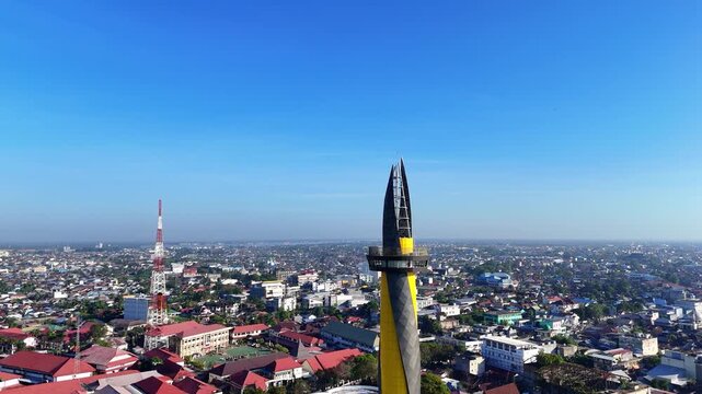 Aerial view of Monument of Pal 0 Banua surrounded by dense urban rooftops and a communication tower under a clear blue sky in Kecamatan Banjarmasin Tengah, Kalimantan Selatan, Indonesia.