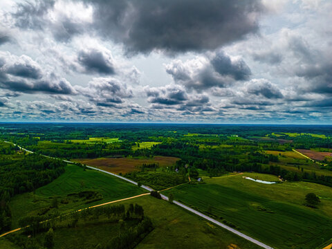 Aerial view of lush green fields, dense forests, and a winding road under a dramatic cloudy sky in Vidzeme, Latvia.