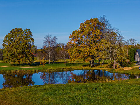 Aerial view of a serene pond reflecting vibrant autumn trees in a lush green landscape under a clear blue sky in Vidzeme, Latvia.