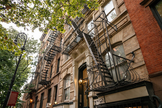 Historic New York brownstone apartment building with classic brick facade and black metal fire escapes viewed from street level under green trees. Urban city architecture with detailed windows