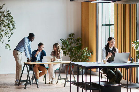 Diverse business team collaborating in a modern office space, smiling colleagues discussing work on laptops, and a woman participating in a video call