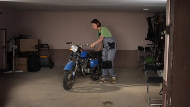 Woman wearing overalls, standing next to a classic blue motorcycle, focusing on its engine in a dimly lit home garage, symbolizing independence, hobby, and hands on work