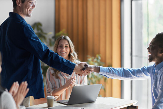 Diverse business people shaking hands, concluding successful partnership deal, celebrating agreement and teamwork in modern office meeting with colleagues applauding