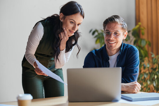 Business colleagues collaborating on a laptop, a man smiling while working, a woman holding papers, discussing project ideas and sharing information in a modern office