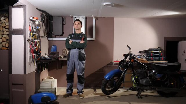 Man mechanic standing confidently with arms crossed, holding wrenches, in a garage workshop filled with various tools, equipment, and a vintage motorcycle, ready for maintenance or repair work