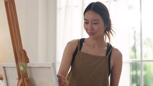 Creative young Asian woman artist painting on canvas in bright home studio. Relaxed female hobbyist sitting on floor, enjoying peaceful indoor leisure time and expressing imagination.