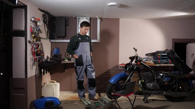Mechanic standing in a garage, looking down at an open toolbox and a laptop while working on a disassembled motorcycle, focusing on the mechanical problem