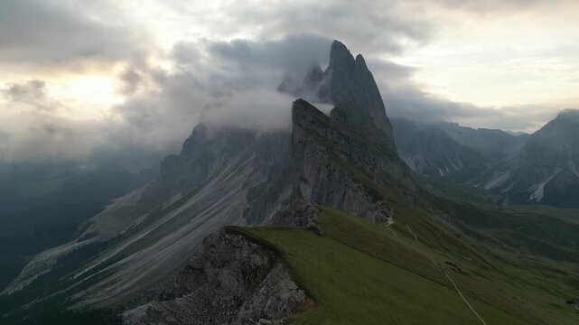 Aerial view of the dramatic Seceda mountain ridge with jagged peaks, grassy slopes, and swirling clouds in the Dolomites, Italy.
