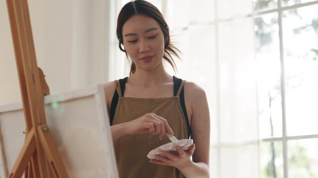 Creative young Asian woman artist painting on canvas in bright home studio. Relaxed female hobbyist sitting on floor, enjoying peaceful indoor leisure time and expressing imagination.