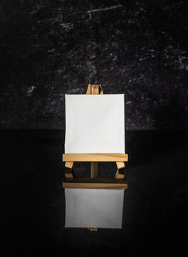 Vertical photo mock up of small square blank white canvas sitting on a mini wooden art easel sitting on black mirror reflective surface and dark background.