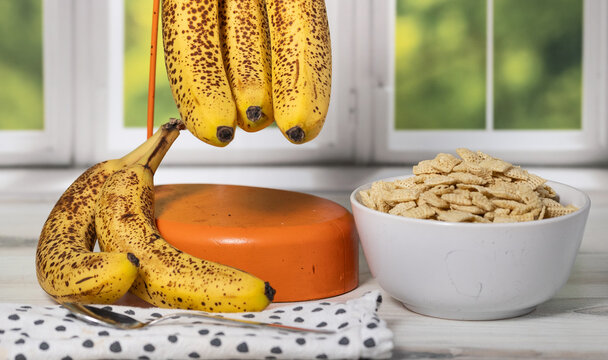 High resolution close up stock photo of sweet ripe brown spotted bananas and full bowl of cereal in front of a kitchen window. 