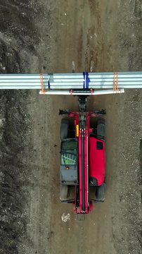 Aerial view of a red telehandler transporting long metal beams along a dirt road in Latvia.