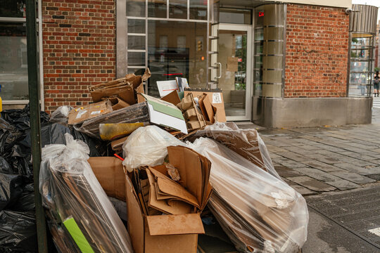Stack of cardboard boxes and plastic trash bags placed on a New York City sidewalk near a brick building entrance. Urban waste management scene highlighting recycling, garbage collection, and street