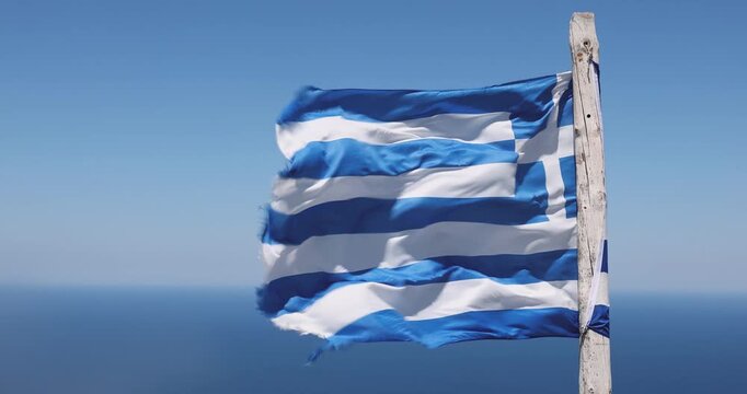 Greek flag on a sun‑bleached wooden stick whips in fierce wind high above the Aegean Sea on Crete, with bright blue water and rugged coastal terrain visible as the camera captures the dramatic motion.