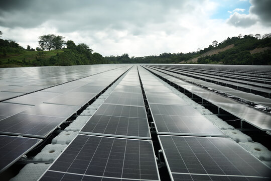 Aerial view of a large floating solar farm on a calm reservoir surrounded by lush green hills under a cloudy sky in Berau Regency, East Kalimantan, Indonesia.